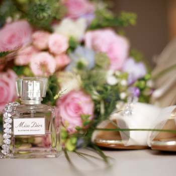 A bride's bouquet lies next to their wedding shoes in a bokeh background. In the foreground, a 'Miss Dior' perfume bottle stands as the image's focal point.