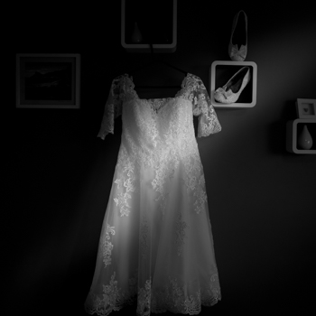 A bride's dress is suspended against a wall with three square shelving units surrounding it. The bride's shoes can be seen in one of the shelving units. The scene is in black and white and is very dark apart from a single beam of natural sunlight illuminating from the top right to bottom left of the dress/scene.
