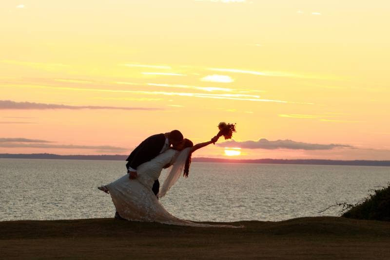 A bride and groom walk hand in hand along a coastal pathway. In the background behind the couple is an epic beach sunset signature to the world famous Swansea Bay. 