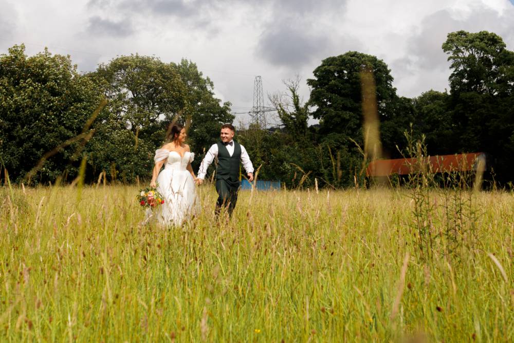 Standing on a hillside on a glorious summer's day a bride and groom can be seen holding hands whilst looking into each other's eyes. Long grass surrounds them whilst the rolling hills of the Welsh countryside can be seen in the scene's background.