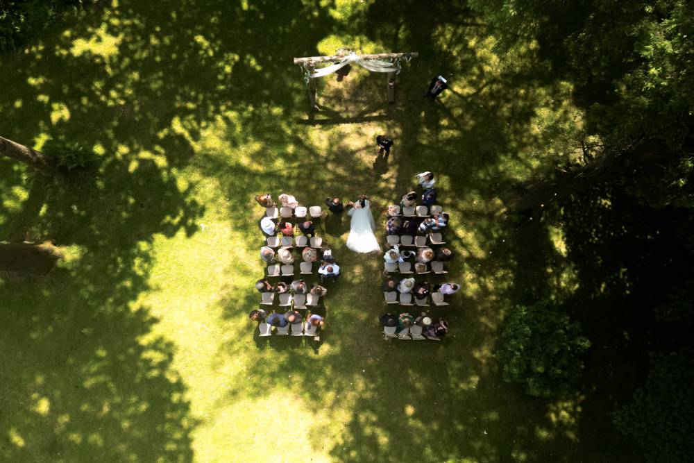In a Welsh woodland setting, two brides can be seen embracing on a small wooden bridge. One brides dress drapes over the bridge while the other bride is wearing a suit. The image is in black and white.