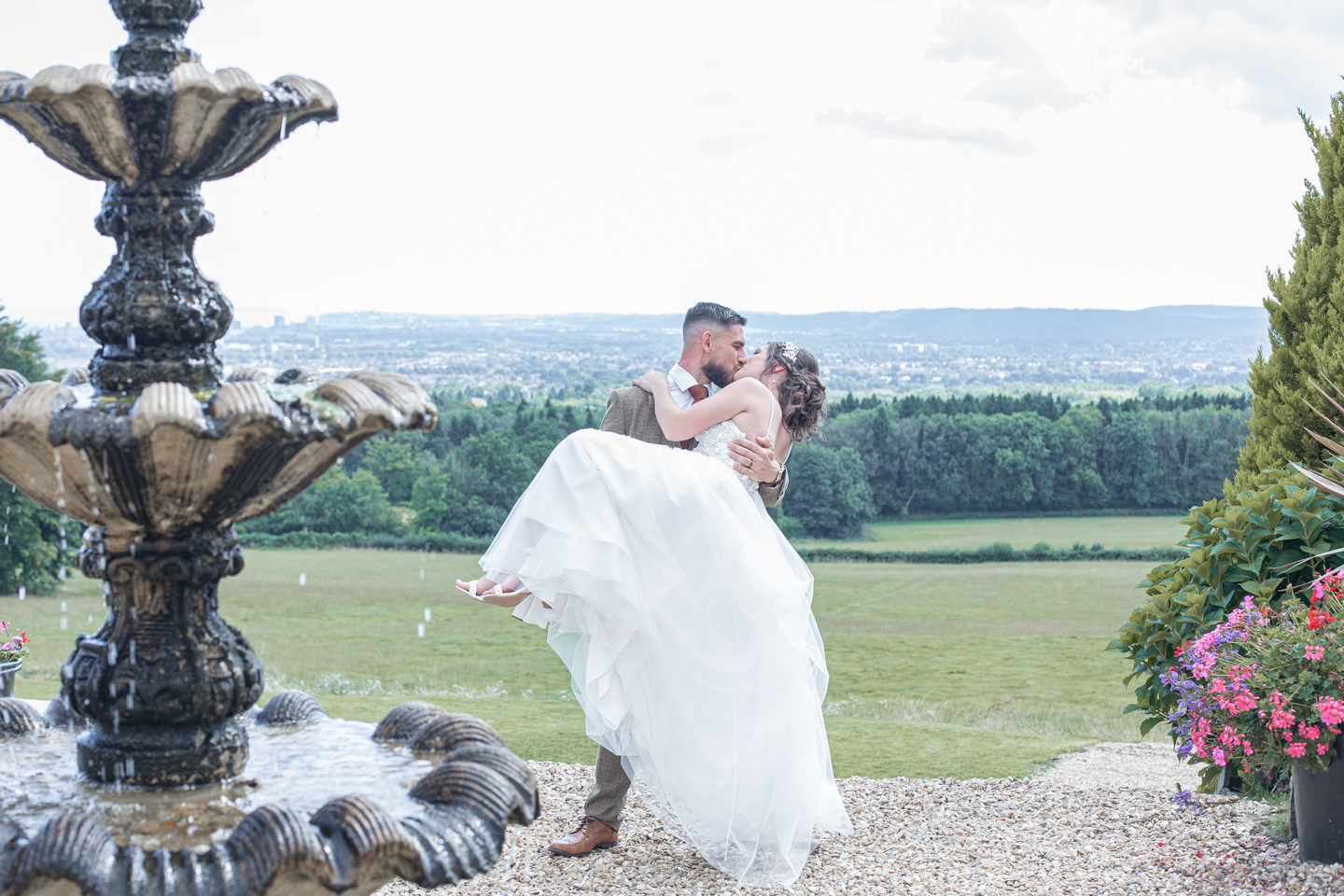 In a Welsh woodland setting, two brides can be seen embracing on a small wooden bridge. One brides dress drapes over the bridge while the other bride is wearing a suit. The image is in black and white.