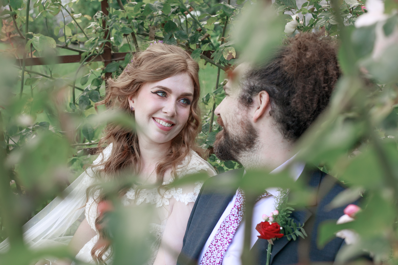 A bride and groom are sat on a bench in a floral bagoda having just married. They can both be seen to smile while looking at one and another.