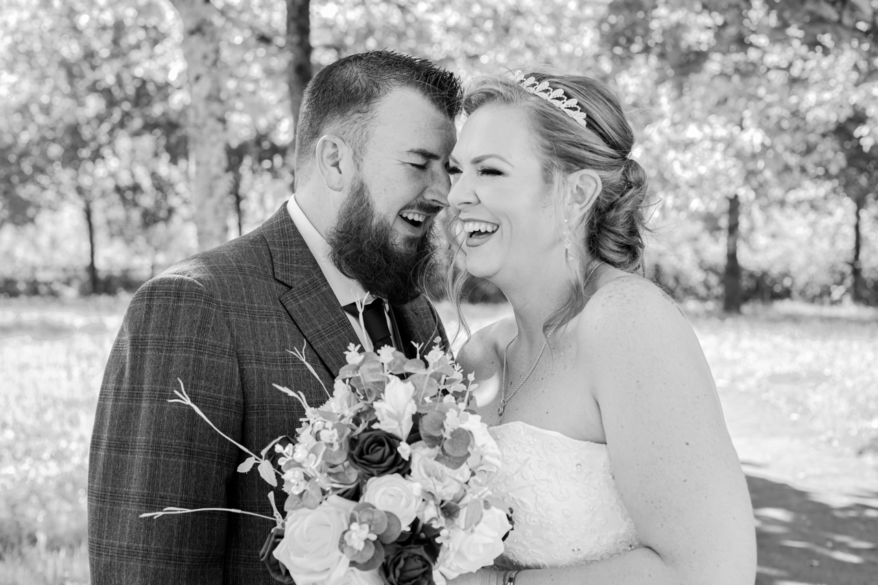 A couple can be seen sharing a laugh on their big day. The bride is holding a bouquet of flowers. The image has been greyscaled into black and white only.