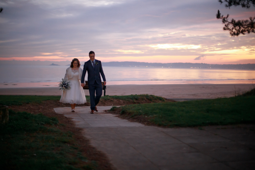 A bride and groom walk hand in hand along a coastal pathway. In the background behind the couple is an epic beach sunset signature to the world famous Swansea Bay. 