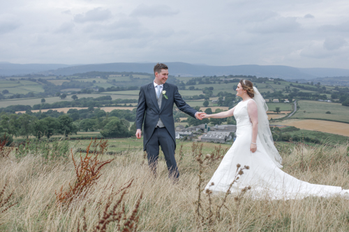 Standing on a hillside on a glorious summer's day a bride and groom can be seen holding hands whilst looking into each other's eyes. Long grass surrounds them whilst the rolling hills of the Welsh countryside can be seen in the scene's background.